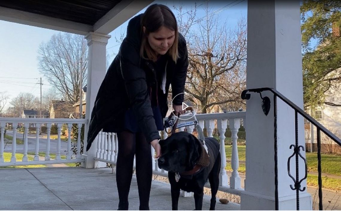 Gabriella pets her black Lab guide dog, Freesia while standing on her house porch.