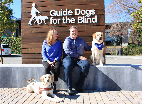 Elizabeth and Tom sit in the courtyard on the GDB campus flanked by two yellow Lab guide dogs.