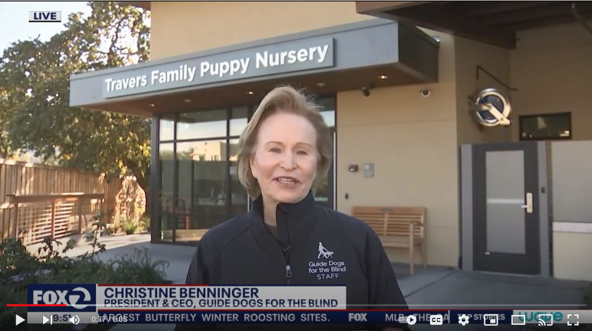 A video screen shot of Christine Benninger standing in front of the Travers Family Puppy Nursery on the GDB California campus.